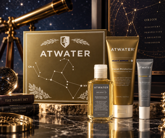 A curated skincare gift set from ATWATER arranged on a polished marble surface with a starry night-sky backdrop and celestial props. The set includes a large gold constellation-themed box, a gold facial moisturizer tube labeled Heavy Armor, a small amber bottle of facial cleansing balm, a gray tube labeled Advanced Repair, and a gray tube labeled Advanced Repair Moisturizer.