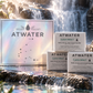 Three ATWATER Clean Impact body scrub and cleansing bars are arranged in front of their iridescent box on a rocky stream with a waterfall in the background. The packaging is holographic with the brand name ATWATER and a constellation graphic, and the bars are white with black text.