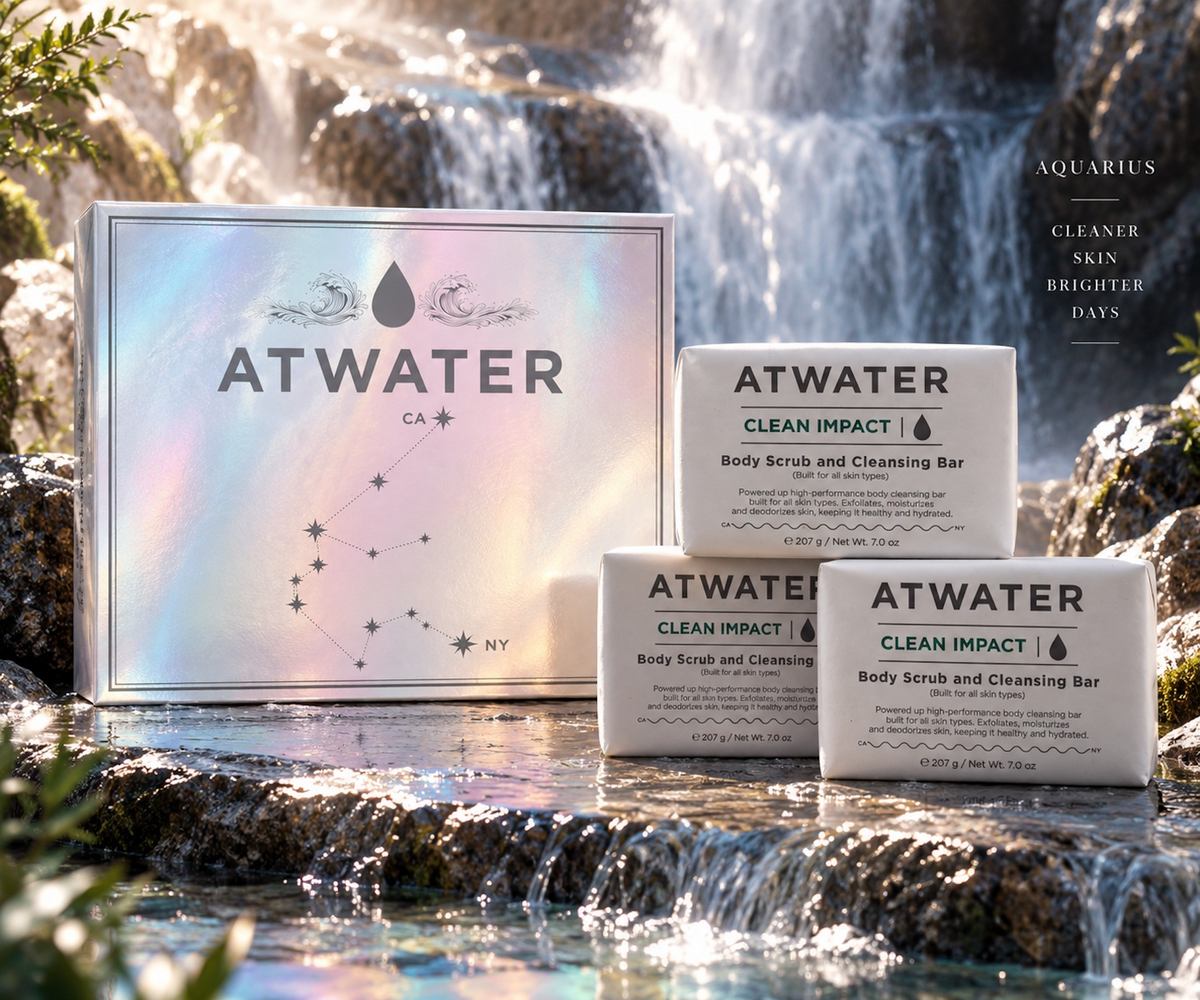 Three ATWATER Clean Impact body scrub and cleansing bars are arranged in front of their iridescent box on a rocky stream with a waterfall in the background. The packaging is holographic with the brand name ATWATER and a constellation graphic, and the bars are white with black text.
