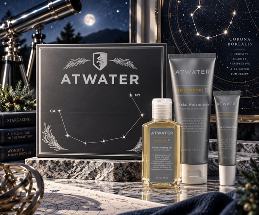 A set of ATWATER skincare products arranged on a marble surface with a dark starry sky and celestial props in the background. The set includes a black gift box with constellation artwork, a clear bottle of facial oil with pale yellow liquid, a gray tube of facial moisturizer, and a gray compact powder tube.