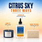 Three ATWater body/candle products arranged on a white surface against a blue-to-yellow gradient background. Text labels identify the set as Citrus Sky Three Ways, with a dark blue travel candle, a clear bottle of hydrating body wash, and a beige bar of body bar.