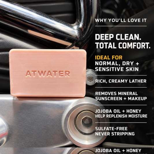 A peach-colored ATWATER soap bar resting on a shiny chrome motorcycle engine, with promotional text on the right detailing its deep clean, creamy lather, and jojoba oil and honey ingredients.