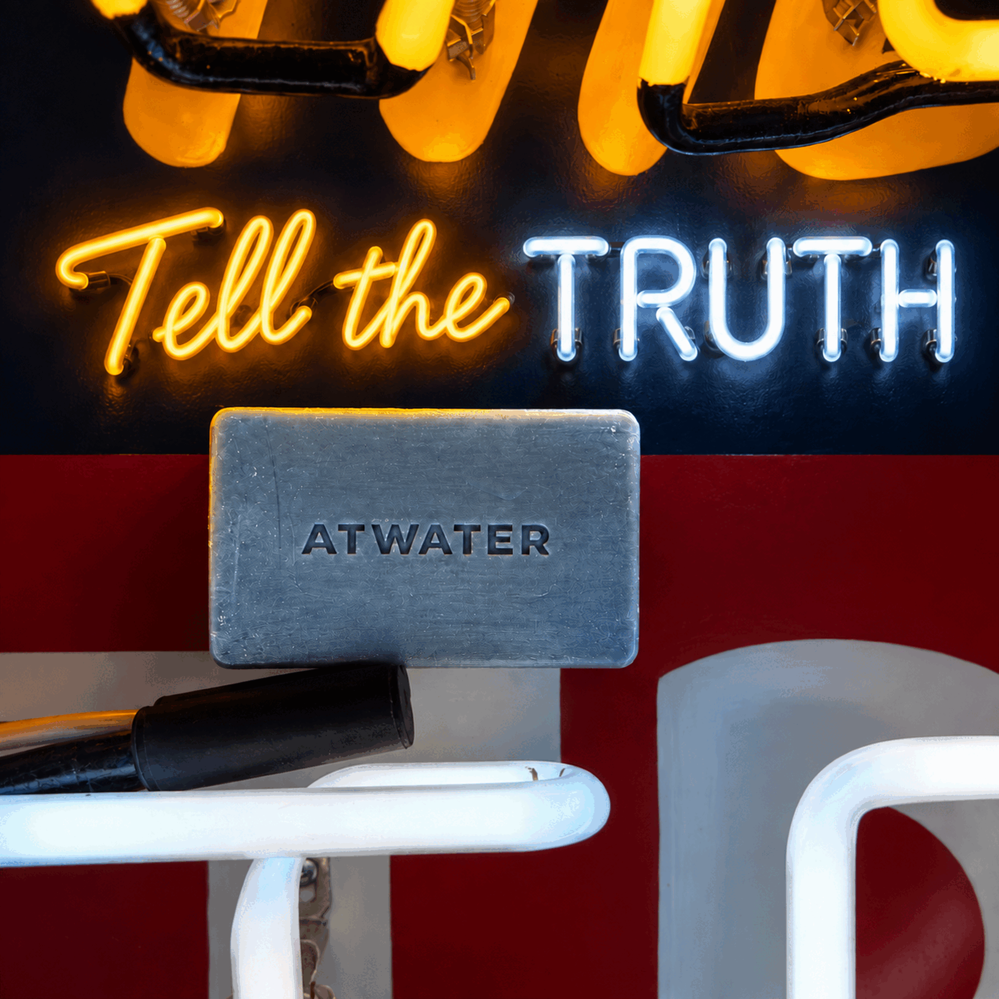 A rectangular gray soap bar embossed with the brand name 'ATWATER' sits on a white metal shelf. The background features a red wall, a black neon sign that reads 'Tell the TRUTH' in yellow and white, and a yellow neon chain link.